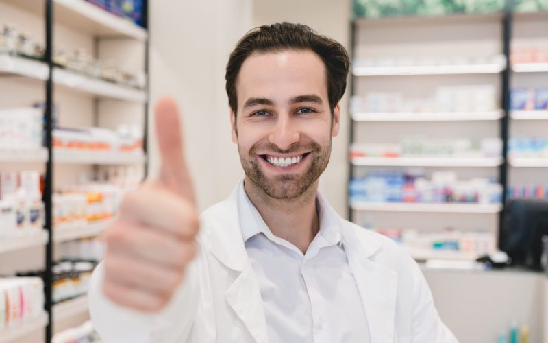 Closeup cropped focused shot of male young caucasian druggist pharmacist showing thumb up in camera in white medical coat in pharmacy drugstore